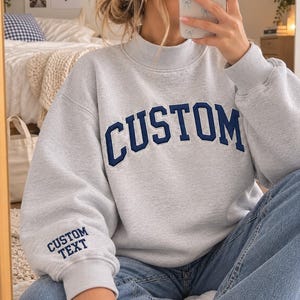 May include: A light gray crewneck sweatshirt with a mock neck collar, featuring the word CUSTOM in dark blue block letters across the chest and the words CUSTOM TEXT embroidered in dark blue on the right sleeve cuff.