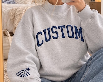 May include: A light gray crewneck sweatshirt with a mock neck collar, featuring the word CUSTOM in dark blue block letters across the chest and the words CUSTOM TEXT embroidered in dark blue on the right sleeve cuff.