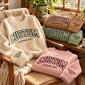 May include: Three knit sweaters in cream, sage green, and dusty pink. Each sweater features a crew neckline, ribbed cuffs, and the word CUSTOM in large block letters with the phrase CUSTOM TEXT underneath. The sleeve cuffs also feature the text CUSTOM TEXT.