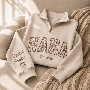 May include: A beige quarter-zip sweatshirt featuring the word NANA in floral-patterned applique letters across the chest, with EST. 2026 embroidered below. The sleeve is embroidered with the names Daniel and Sophia above two small hearts.