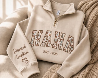 May include: A beige quarter-zip sweatshirt featuring the word NANA in floral-patterned applique letters across the chest, with EST. 2026 embroidered below. The sleeve is embroidered with the names Daniel and Sophia above two small hearts.