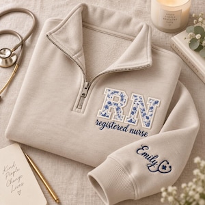 May include: A beige quarter-zip sweatshirt featuring the letters RN in a blue floral pattern with the text registered nurse embroidered below. The sleeve displays the name Emily C with a stethoscope icon. The fabric has a soft, fleece-like texture.