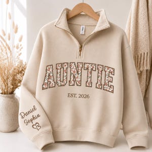May include: A beige quarter-zip sweatshirt featuring the word AUNTIE in large block letters with a floral pattern. Below the text is EST. 2026. The sleeve is embroidered with the names Daniel and Sophia above a small heart icon.
