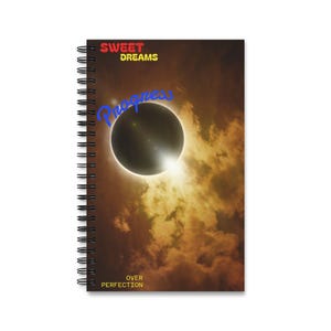 Puede incluir: Un cuaderno de espiral con un gráfico de eclipse solar sobre un cielo nublado de color marrón dorado. El texto "SWEET DREAMS" aparece en rojo y amarillo, "Progress" en azul y "OVER PERFECTION" en amarillo en la portada.
