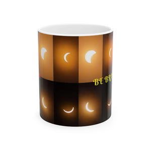 Puede incluir: Una taza de cerámica blanca con un patrón repetitivo de fases lunares en cuarto creciente sobre un fondo degradado de tonos marrón oscuro y ámbar dorado. El texto BE BRAVE está impreso en amarillo en el lateral de la taza, sobre el fondo oscuro.