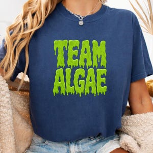 Peut inclure: Un t-shirt bleu marine à manches courtes avec le texte "TEAM ALGAE" imprimé en vert citron vif dans une police dégoulinante rappelant de la vase sur la poitrine. Le vêtement présente un col rond et une coupe standard en tissu bleu foncé.