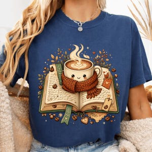Peut inclure: Un t-shirt bleu marine avec une illustration d'un livre ouvert sur lequel repose une tasse de café fumante portant une écharpe en tricot marron. Le motif comprend des grains de café, des petites feuilles et des éléments décoratifs autour du livre.