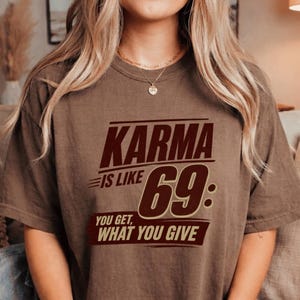 May include: A brown short-sleeve t-shirt featuring dark brown and cream text that reads: KARMA IS LIKE 69: YOU GET, WHAT YOU GIVE. The shirt has a crew neckline and a relaxed fit, with the graphic centred prominently on the front chest area.