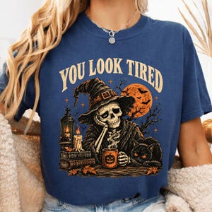 Peut inclure: Un t-shirt bleu marine avec un motif représentant un squelette portant un chapeau de sorcière, la tête appuyée sur une main. Le squelette est assis près d'une lanterne, de livres empilés et d'un chat noir, tenant une tasse citrouille.