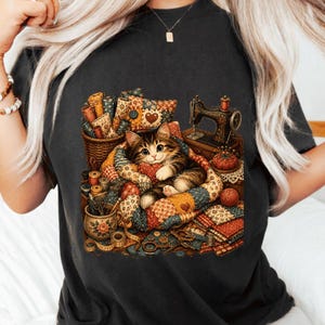 以下が含まれることがあります： カラフルなパッチワークキルトの中で丸まっている茶色のトラ猫のグラフィックイラストが描かれたダークグレーのTシャツ。ヴィンテージのミシン、布ロールが入ったバスケット、糸巻き、ハサミ、花柄のマグカップが描かれています。
