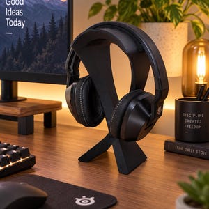 Op de afbeelding: Een zwarte over-ear hoofdtelefoon op een zwarte X-vormige hoofdtelefoonstandaard. De hoofdtelefoon heeft een gevoerde hoofdband en oorschelpen met zichtbare stiksels. De standaard heeft een brede voet en een gebogen steun voor de hoofdtelefoon.