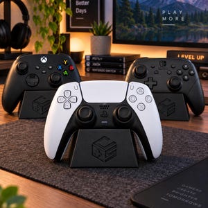 Op de afbeelding: Drie zwarte plastic gamecontroller-standaards, elk met een geometrisch logo op de voorzijde. De standaards houden een zwarte Xbox-controller, een zwarte Nintendo Switch Pro-controller en een wit-zwarte PlayStation 5 DualSense-controller vast.