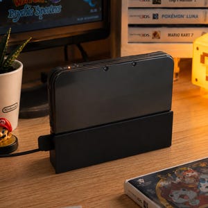 May include: A black handheld gaming console docked in a black charging cradle. The console is closed, featuring a small orange indicator light on the top edge. A black power cable extends from the side of the charging base, which sits on a wooden surface.