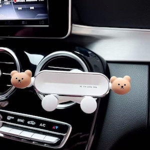 May include: A white car air vent clip featuring a horizontal rectangular frame with rounded edges, two light brown bear heads on the sides, and two white circular knobs at the bottom. The text "Hi I'm Little One" is printed on the white surface.