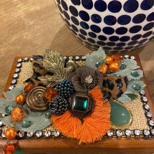 May include: A rectangular wooden box with a burlap top surface, decorated with a cluster of orange and green beads, a bronze spiral, a dark green gemstone, an orange tassel, leopard print fabric, and a border of small clear rhinestones.