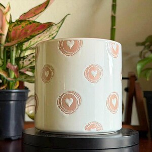 May include: A cylindrical white ceramic planter featuring a repeating pattern of embossed dusty pink circles, each containing a small white heart at its center. The surface has a smooth, glossy finish with evenly spaced circular motifs throughout.