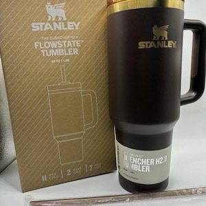 Puede incluir: Un vaso Stanley Quencher H2.0 FlowState de color marr&oacute;n oscuro con capacidad de 1,18 L, borde dorado y asa a juego. Se muestra junto a su embalaje de cart&oacute;n original y una pajita envuelta en pl&aacute;stico sobre una superficie de color blanco.