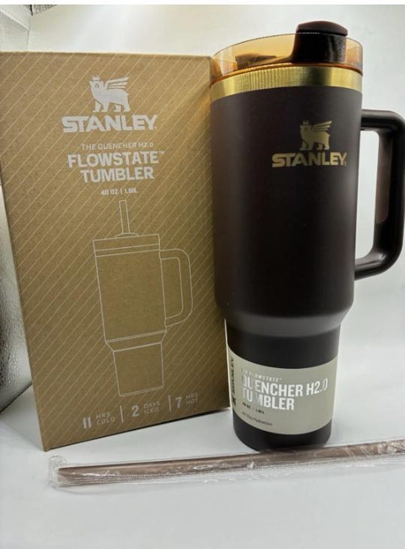 Puede incluir: Un vaso Stanley Quencher H2.0 FlowState de color marr&oacute;n oscuro con capacidad de 1,18 L, borde dorado y asa a juego. Se muestra junto a su embalaje de cart&oacute;n original y una pajita envuelta en pl&aacute;stico sobre una superficie de color blanco.