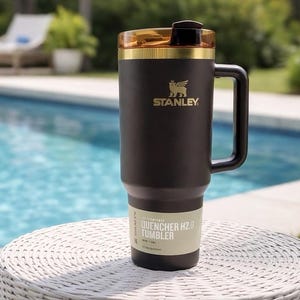 Puede incluir: Un vaso Stanley Quencher H2.0 de color marr&oacute;n oscuro mate con un borde dorado y una tapa &aacute;mbar transl&uacute;cida. El vaso cuenta con un asa lateral, un logotipo de Stanley dorado y una etiqueta en la base que dice Quencher H2.0 Tumbler 40 oz 1,18 L.