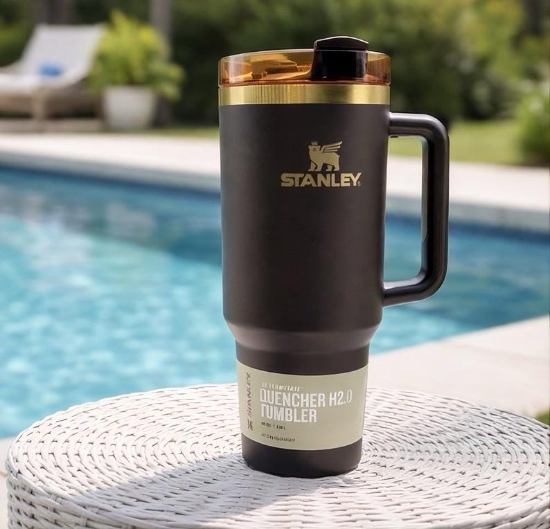 Puede incluir: Un vaso Stanley Quencher H2.0 de color marr&oacute;n oscuro mate con un borde dorado y una tapa &aacute;mbar transl&uacute;cida. El vaso cuenta con un asa lateral, un logotipo de Stanley dorado y una etiqueta en la base que dice Quencher H2.0 Tumbler 40 oz 1,18 L.
