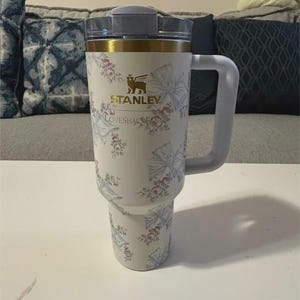 Może przedstawiać: Biały kubek Stanley z uchwytem, ozdobiony złotym brzegiem oraz wzorem w kwiaty i wstążki. Z przodu nadrukowano napisy Stanley oraz LoveShackFancy. Kubek posiada przezroczystą plastikową pokrywkę i zwężaną podstawę pasującą do uchwyt&oacute;w na napoje.