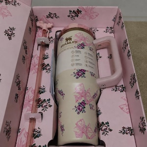 Puede incluir: Un vaso Stanley de color crema con un patr&oacute;n floral y de lazos rosas, que cuenta con un asa rosa a juego y una pajita larga de color rosa transl&uacute;cido. El vaso se presenta en una caja rosa a juego decorada con un estampado floral y de lazos.