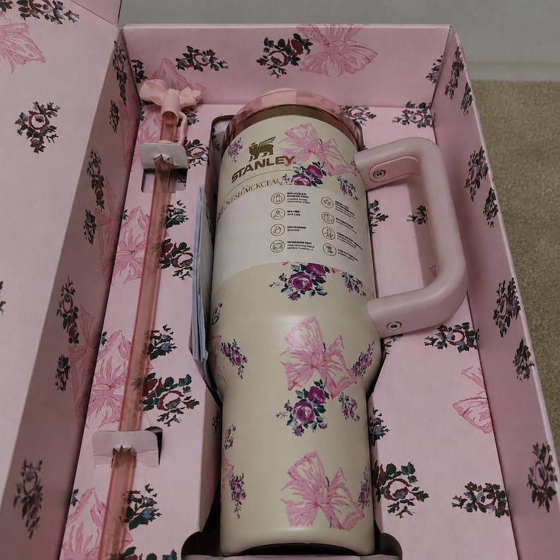Puede incluir: Un vaso Stanley de color crema con un patr&oacute;n floral y de lazos rosas, que cuenta con un asa rosa a juego y una pajita larga de color rosa transl&uacute;cido. El vaso se presenta en una caja rosa a juego decorada con un estampado floral y de lazos.