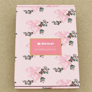 Puede incluir: Una caja rectangular de cart&oacute;n rosa con un patr&oacute;n repetitivo de lazos rosa claro y peque&ntilde;os ramos de flores. Centrada en la parte frontal hay una etiqueta rectangular con borde dorado que contiene el texto blanco Stanley y LoveShackFancy.