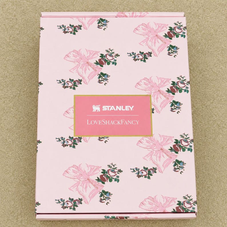 Puede incluir: Una caja rectangular de cart&oacute;n rosa con un patr&oacute;n repetitivo de lazos rosa claro y peque&ntilde;os ramos de flores. Centrada en la parte frontal hay una etiqueta rectangular con borde dorado que contiene el texto blanco Stanley y LoveShackFancy.