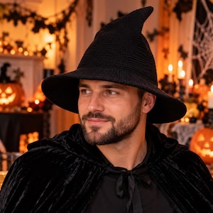 May include: A black knitted witch hat with a pointed crown and a wide, slightly downturned brim. The hat features a textured, woven fabric construction and is worn with a black velvet-textured cape that ties at the neck with a fabric ribbon.