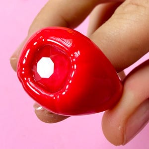 Könnte beinhalten: Ein klobiger, glänzender roter Ring mit einem glatten, abgerundeten Band und einem mittig eingesetzten, facettierten, klaren achteckigen Stein. Der Ring hat ein kräftiges, massives Erscheinungsbild mit einer hochglänzenden Oberfläche, die das Licht reflektiert.