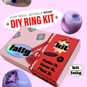 Op de afbeelding: Een DIY-ringset in een rechthoekige doos met roze en rode panelen. De doos bevat tekst zoals A DIY YOU'LL ACTUALLY WEAR!, DIY RING KIT, fatty ring en Make it. Wear it. Flex it. Twee dikke ringen met ingebedde kristallen zijn afgebeeld.