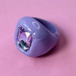 Puede incluir: Un anillo grueso de color lavanda con una banda lisa y redondeada y una gran piedra preciosa morada de corte cuadrado y múltiples facetas engastada en el centro. El anillo tiene un acabado brillante y sólido que contrasta con el cristal claro.
