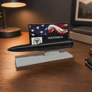May include: A black bullet-shaped business card holder mounted on a rectangular light gray base. The holder features a slot containing a card with an American flag graphic and the text Tactical Display Co. printed in white on a black background.