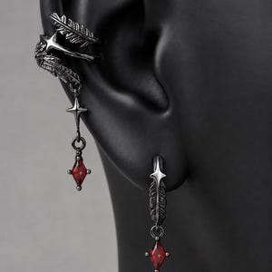 May include: A dark metal ear cuff and earring set featuring detailed feather motifs. The ear cuff has a dangling star charm and a red diamond-shaped crystal. The matching earring features a feather design with a dangling red diamond-shaped crystal.