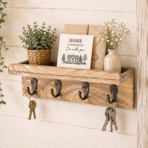 May include: A wooden wall-mounted shelf with four dark metal hooks. The shelf holds a small potted plant in a woven basket, a tan envelope, and a white sign with the text "HOME IS WHEREVER I'm with you." and a house illustration. Keys hang from two hooks.