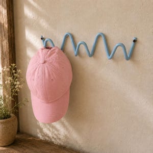 May include: A light blue wall-mounted coat rack with a continuous wavy, zigzag design. A solid pink baseball cap with a structured crown and a curved brim is hanging from the first loop of the metal rack, which is secured to the wall with screws.
