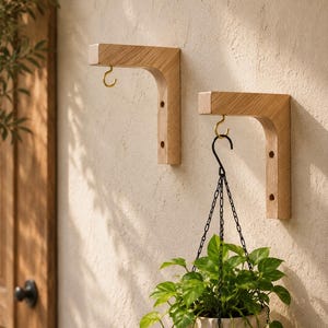May include: Two L-shaped light wood wall brackets with a natural grain finish. Each bracket features a small gold-toned metal hook attached to the underside. One bracket supports a hanging plant in a white textured pot suspended by a black chain.