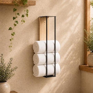 May include: A wall-mounted towel rack featuring a vertical light wood back panel and a black metal frame. The rack holds three neatly rolled white towels stacked vertically within the black metal structure, providing a storage solution.