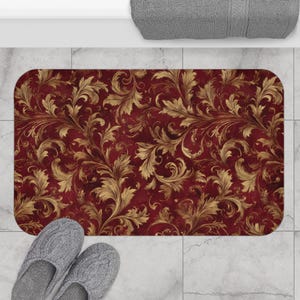 May include: A rectangular bath mat featuring a deep burgundy background with a repeating gold-toned acanthus leaf scroll pattern. The mat has rounded corners and is positioned on a light gray tiled floor with a pair of gray slippers nearby.