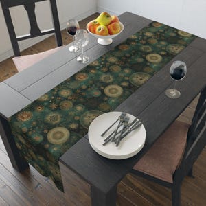 May include: A dark green table runner featuring a repeating pattern of golden circular mandala-like designs and starburst motifs. The runner is laid across a dark wooden dining table set with a stack of white plates, silverware, and wine glasses.