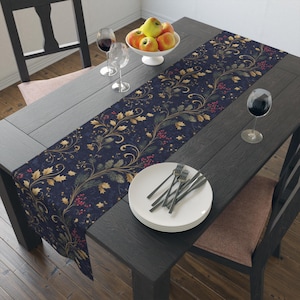 May include: A dark blue table runner featuring a gold and green botanical pattern with red berries, laid across a dark wooden table with a stack of white plates, three wine glasses, and a white bowl filled with red and yellow apples.
