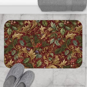 May include: A rectangular bath mat featuring a deep red background with a repeating pattern of gold acanthus leaves, green pine needles, holly leaves, and clusters of red berries. The mat has rounded corners and a soft, textured surface.
