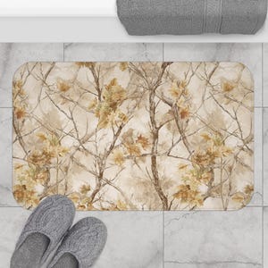 May include: A rectangular bath mat featuring a detailed camouflage pattern of brown and tan tree branches with autumn leaves. The mat has rounded corners and is positioned on a light grey tiled floor with a pair of grey slippers nearby.