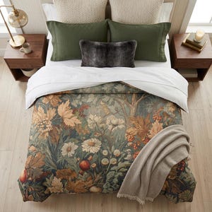 Op de afbeelding: Een bed met een dekbedovertrek met een botanische print van herfstbladeren, bessen en witte bloemen in tinten groen, oranje en bruin. Het bed bevat twee olijfgroene kussenslopen, twee crèmekleurige kussens met textuur en een grijs imitatiebont kussen.
