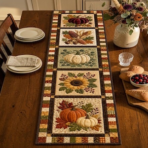 May include: A rectangular fabric table runner featuring a patchwork design with five distinct square panels. Each panel displays seasonal autumn motifs including pumpkins, corn, sunflowers, and fall leaves in shades of orange, yellow, and green.