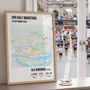 Könnte beinhalten: Ein gerahmtes Poster mit einer Karte der CPH Half Marathon Strecke und dem Text CPH HALF MARATHON 20 SEPTEMBER 2026, OLA NORDMAN #0182, 21,09 KM, 01:26 und 4:55/KM. Eine goldfarbene Medaille liegt auf der Oberfläche neben dem Holzrahmen.