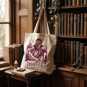 Può includere: Una borsa tote in tela beige con una stampa grafica rosso scuro raffigurante un vampiro, un castello, croci e pipistrelli. Il testo "DRACULA BY BRAM STOKER" è stampato con un carattere stilizzato nella parte inferiore del disegno.
