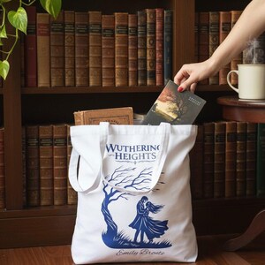 Può includere: Una borsa tote in tela bianca con una stampa grafica blu che raffigura una coppia in piedi sotto un albero. Il testo Wuthering Heights è stampato sopra l'illustrazione e il nome Emily Bronte è riportato sotto il grafico in corsivo.