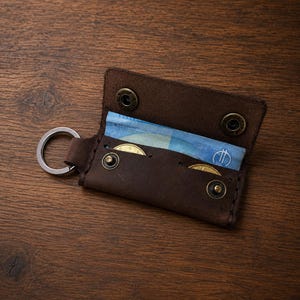Könnte beinhalten: Ein dunkelbrauner Schlüsseletui aus Leder mit einem seitlich befestigten Metallring. Das Etui ist geöffnet und zeigt einen gefalteten blauen Geldschein sowie zwei goldfarbene Münzen im Inneren. Es verfügt über vier Druckknöpfe aus Messing.
